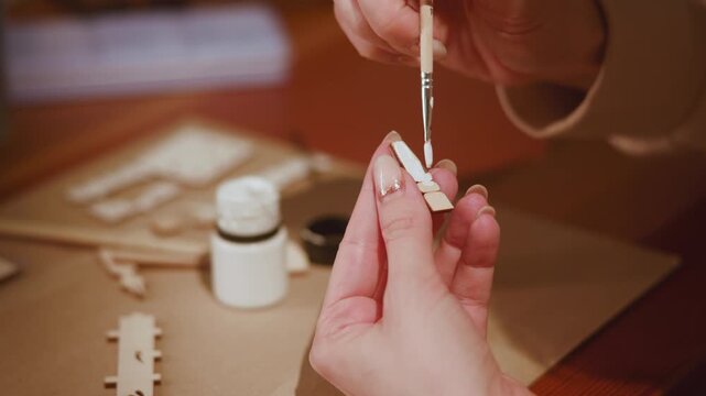 Hands applying white glue with fine paintbrush to small unfinished wooden model piece in warm craft workspace, showing careful hobby assembly, metallic brush ferrule, close detail