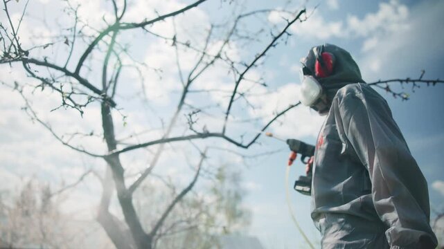 Hooded orchard worker in protective coverall holding handheld sprayer beside bare branches under bright cloudy daylight, standing in calm rural field with safety focused posture