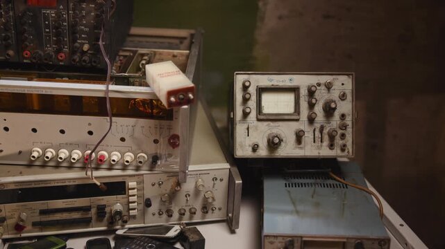 Stacked vintage electronic test equipment with oscilloscope, old mobile phones, cables, knobs, dials, and worn metal parts on cluttered workshop bench in muted industrial light