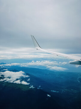 Passenger airplane wing flying high above beautiful blue mountains and white cumulus clouds under a dramatic sky. Scenic view from a plane window during a flight journey.