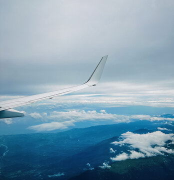 Passenger airplane wing flying high above beautiful blue mountains and white cumulus clouds under a dramatic sky. Scenic view from a plane window during a flight journey.