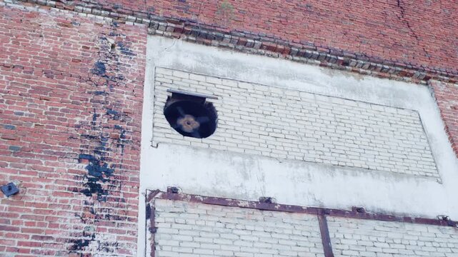 Rusted industrial fan turning inside circular brick opening with whitewashed infill and weathered red facade from low upward viewpoint, showing gritty urban decay texture and detail