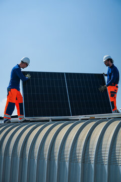 Two renewable energy specialists in hard hats and hi vis workwear steadily carry a single photovoltaic module up the curved roof together high on an industrial warehouse roof in a Thai city.