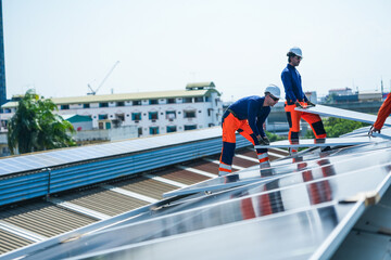 Fototapeta premium A pair of photovoltaic technicians wearing safety vests and hard hats steadily secure the new panel helped by the installation crew on the curved metal roof of an industrial factory.
