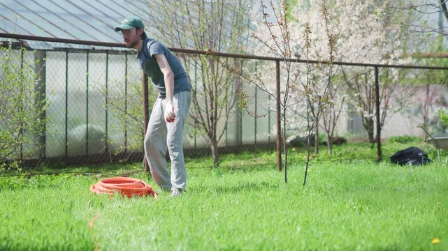 Man bending to pick up red bag on grassy lawn beside metal fence with trees in background, side view in soft daylight, showing outdoor maintenance in calm suburban setting