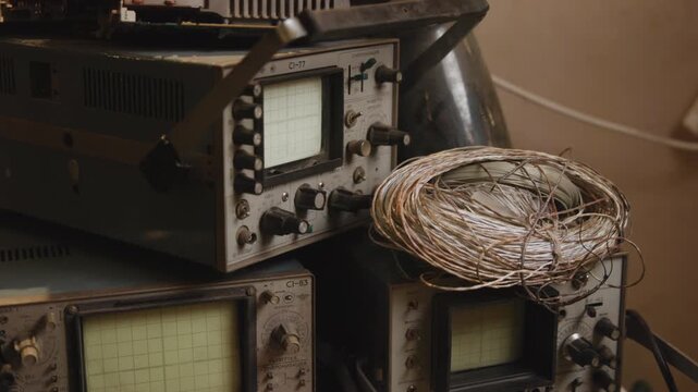 Stacked vintage oscilloscopes and coiled copper wire on workshop bench under warm directional light, showing rotary controls, CRT screens, metal housings, and repair details
