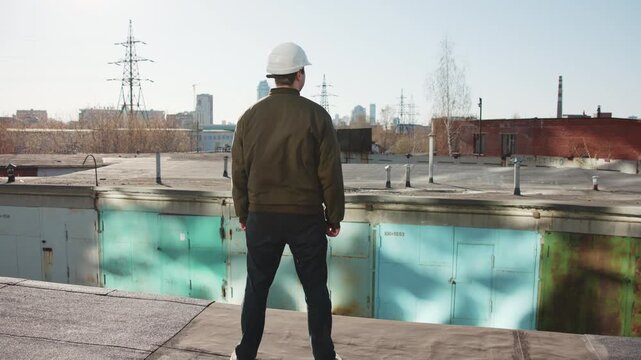 Rear view construction inspector standing on flat industrial rooftop, wearing white hard hat and olive jacket, overlooking urban skyline with concrete surfaces, metal structures, muted daylight