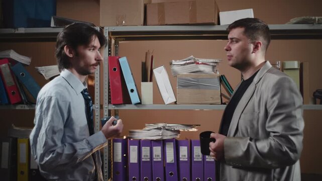 Two office workers in suits face to face in cluttered storage aisle, holding folders beside metal shelves packed with binders, papers, boxes, and files during tense discussion