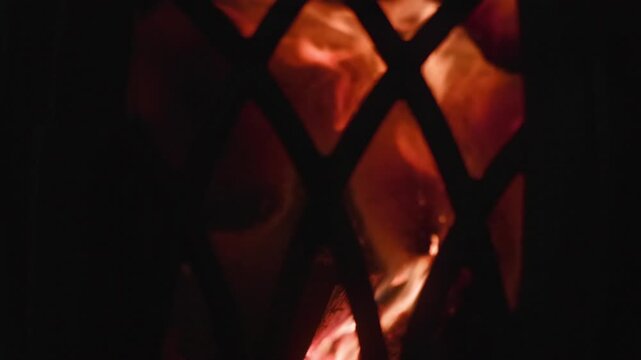 Close-up flames glowing and flickering behind diamond-pattern metal mesh, with warm amber light, deep shadows, and strong contrast in dark moody scene, with fire, flame, ember, glow detail