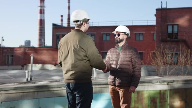 Construction workers shaking hands near red brick industrial building beside water pool, side view under daylight, showing professional agreement, conversation, teamwork, and site collaboration