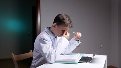 Fototapeta premium Medical professional feeling exhausted and rubbing eyes, sitting at a desk with a laptop and documents, symbolizing the toll of stress and demanding work hours