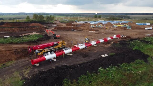 Aerial view of pipeline installation on rural construction site with crews, heavy equipment, concrete supports, long red and white pipe sections, trench prep, metal fittings, overcast daylight, wide