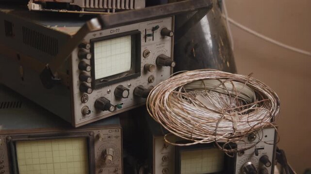 Stacked vintage oscilloscopes beside coiled braided copper wire on workshop shelf, showing aged metal controls, worn surfaces, and industrial texture in warm indoor light