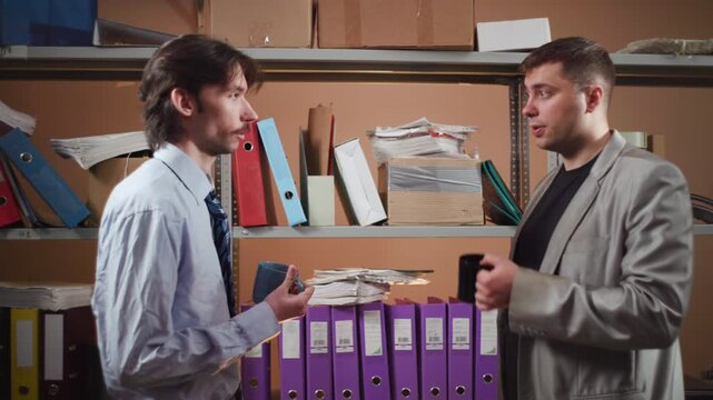 Two coworkers facing each other in office storage room with metal shelving, binders, folders, raised mugs, hand gestures, serious workplace conversation under soft light