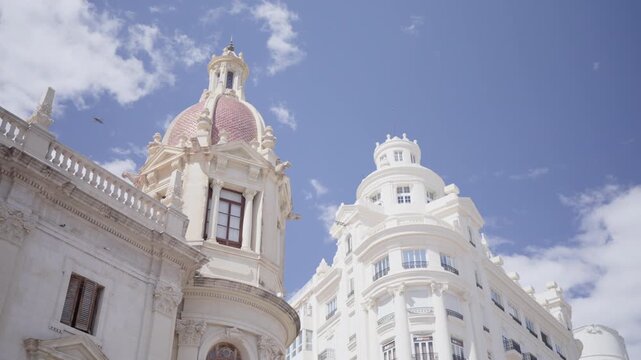 Low angle view of historic white stone buildings with ornate Baroque dome, rounded tower, carved facades, arched windows, and pastel details beneath clear blue sky