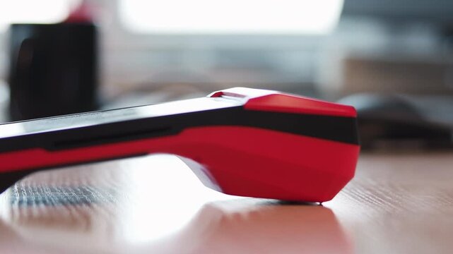 Low angle close-up of red and black handheld device on wooden tabletop with hand reaching in to grasp upper panel during product pickup, with hand, grasp, touch, interaction detail