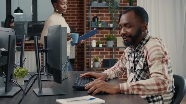 Software developer types programming code on an office keyboard, turning from the monitor during software development work, around a detailed workstation setup.