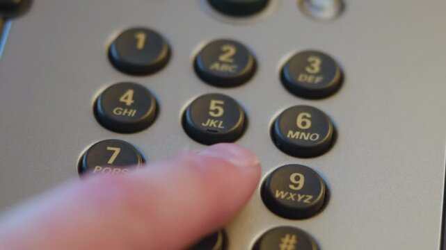 Close-up fingertip pressing numeric keypad on office desk telephone under warm diffuse light, showing tactile contact, recessed round keys, matte silver housing, shallow focus