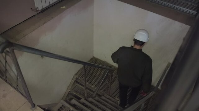 Industrial worker descending narrow metal stairwell in service shaft with hard hat, handrail, grated steps, scuffed wall, muted light, confined maintenance passage