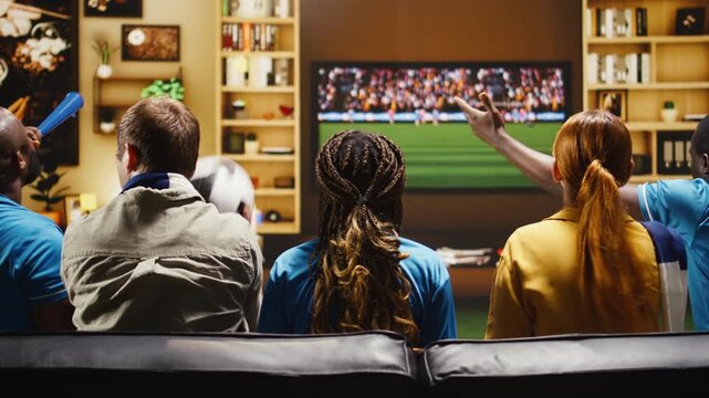 Football supporters eat popcorn and drink beer while watching a live match on TV as friends hold fan props and enjoy snacks during a home watch party.