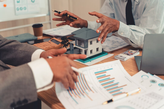 Real estate agent explaining housing details and loan conditions to client with house model on desk.
