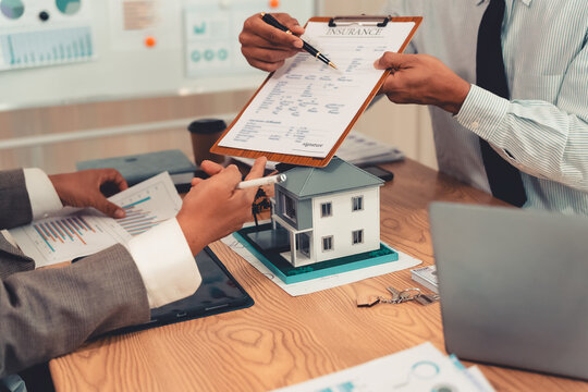 Real estate agent explaining housing details and loan conditions to client with house model on desk.