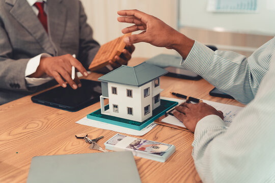 Real estate agent explaining housing details and loan conditions to client with house model on desk.