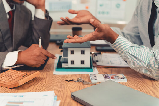 Real estate agent explaining housing details and loan conditions to client with house model on desk.
