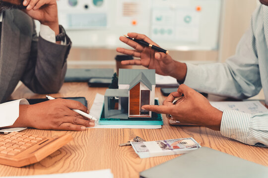 Real estate agent explaining housing details and loan conditions to client with house model on desk.