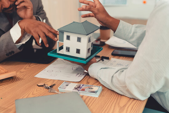 Real estate agent explaining housing details and loan conditions to client with house model on desk.