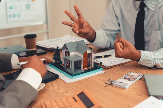 Real estate agent explaining housing details and loan conditions to client with house model on desk.