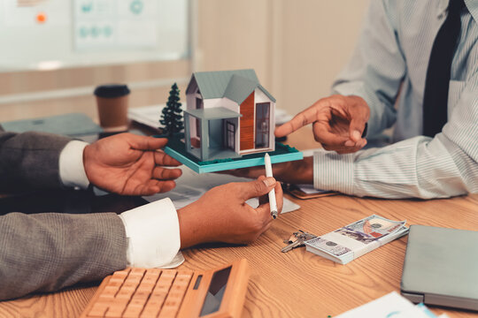 Real estate agent explaining housing details and loan conditions to client with house model on desk.