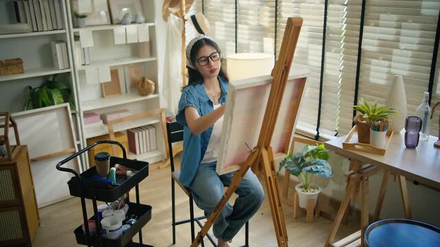 Young Asian woman artist painting on canvas easel with paintbrush in art studio, representing creative professional women designer, fine arts student lifestyle and freelance illustrator