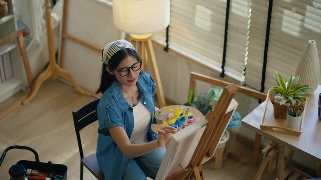 Young Asian woman artist painting on canvas easel with paintbrush in art studio, representing creative professional women designer, fine arts student lifestyle and freelance illustrator