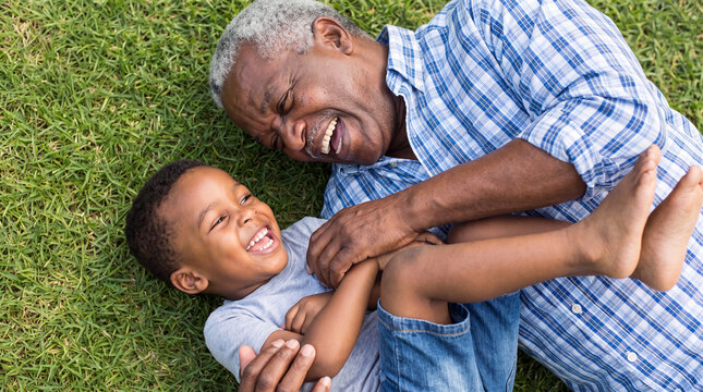 A cheerful African American grandfather tickles and plays with his young grandson while lying on the green grass in a park, capturing a joyful family moment.