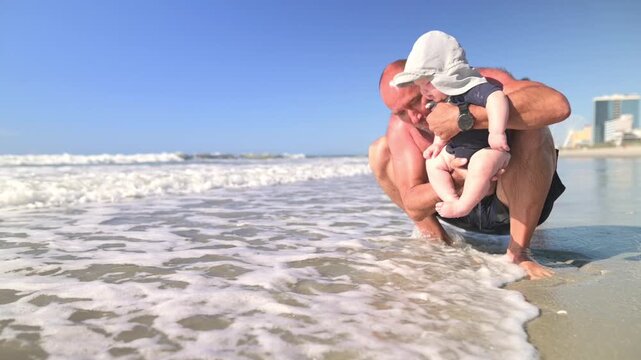 Caucasian man in black swim shorts crouches down to engage with a baby wearing a sun hat at the beach, with soft waves washing over their feet and a bright blue sky above