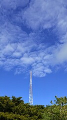 Obraz premium Tall communication tower above green trees under cloudy blue sky