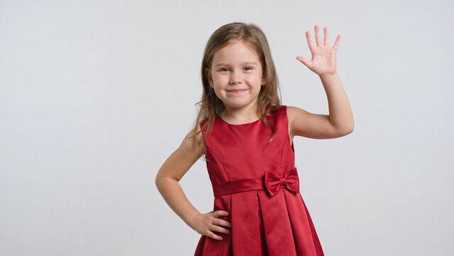 Girl in a red dress waves her hand while smiling confidently against a plain light gray studio background with ample room for text.