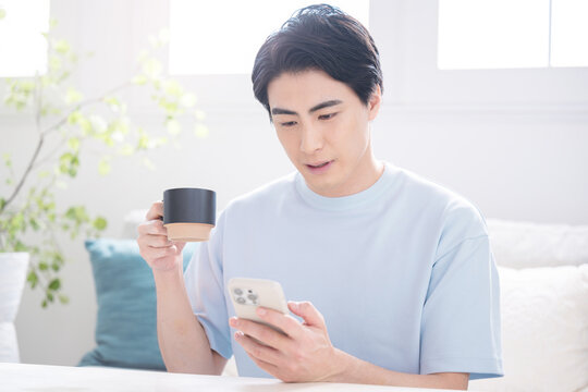 スマートフォンを操作する日本人男性　コーヒーを飲みながら情報を確認する在宅ライフスタイル　スマホを見て驚く