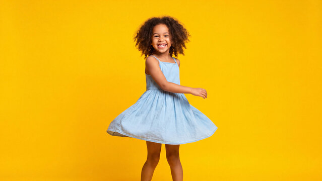 Girl with curly hair dances happily in a light blue dress while spinning against a bright solid yellow background in a studio.