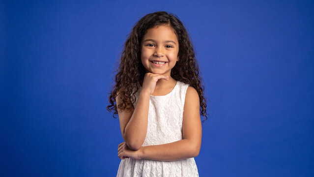 Girl with curly hair wearing a white dress stands against a blue background and rests her chin on her hand while smiling brightly at the camera lens for this studio portrait.