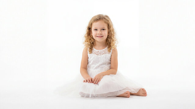 Girl with curly hair sits on the studio floor wearing a formal white dress and looks into the lens with a soft smile while posing in a bright environment suitable for commercial projects.
