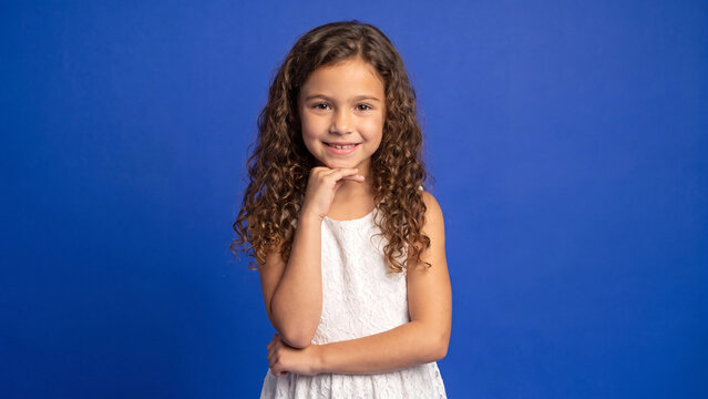 Girl with curly hair wearing a white summer dress poses thoughtfully against a solid blue studio background during a portrait session.