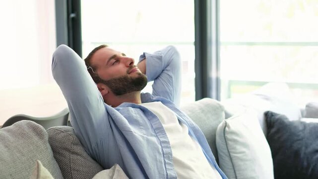 Young handsome man with a beard relaxing on a couch at home, leaning back with hands behind his head and a happy smile, enjoying a moment of tranquility in his bright living room