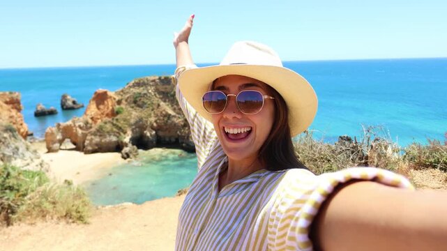 Beautiful smiling woman wearing a hat and sunglasses taking a selfie video from a cliff top viewpoint with an amazing turquoise water beach and rock formations in the background on a sunny day