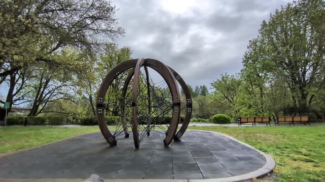 Innovative circular rope climbing playground with intersecting cables and soft rubber safety flooring sits in a peaceful park surrounded by green lawns and benches after spring rain.