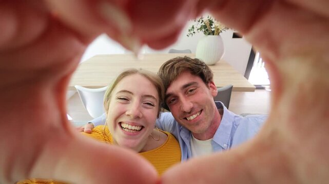 Joyful young couple sitting on a couch, laughing and looking at the camera while making a heart shape with their hands, symbolizing their love, affection, and strong relationship at home