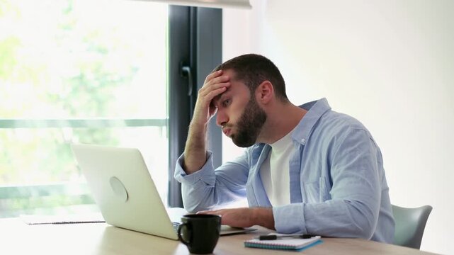 Frustrated young man experiencing professional burnout and headache from overworking on his laptop, feeling stressed and tired while sitting at his desk in a modern home office environment
