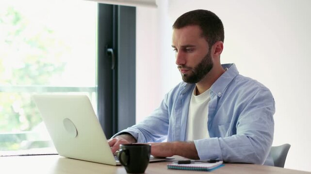 Frustrated young man experiencing headache and fatigue while working on a difficult project at his home office desk, showing signs of burnout from overworking and stress