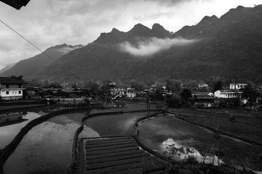 A serene landscape features rice paddies reflecting the moody sky, with mountains shrouded in mist creating a dramatic backdrop - horizontal